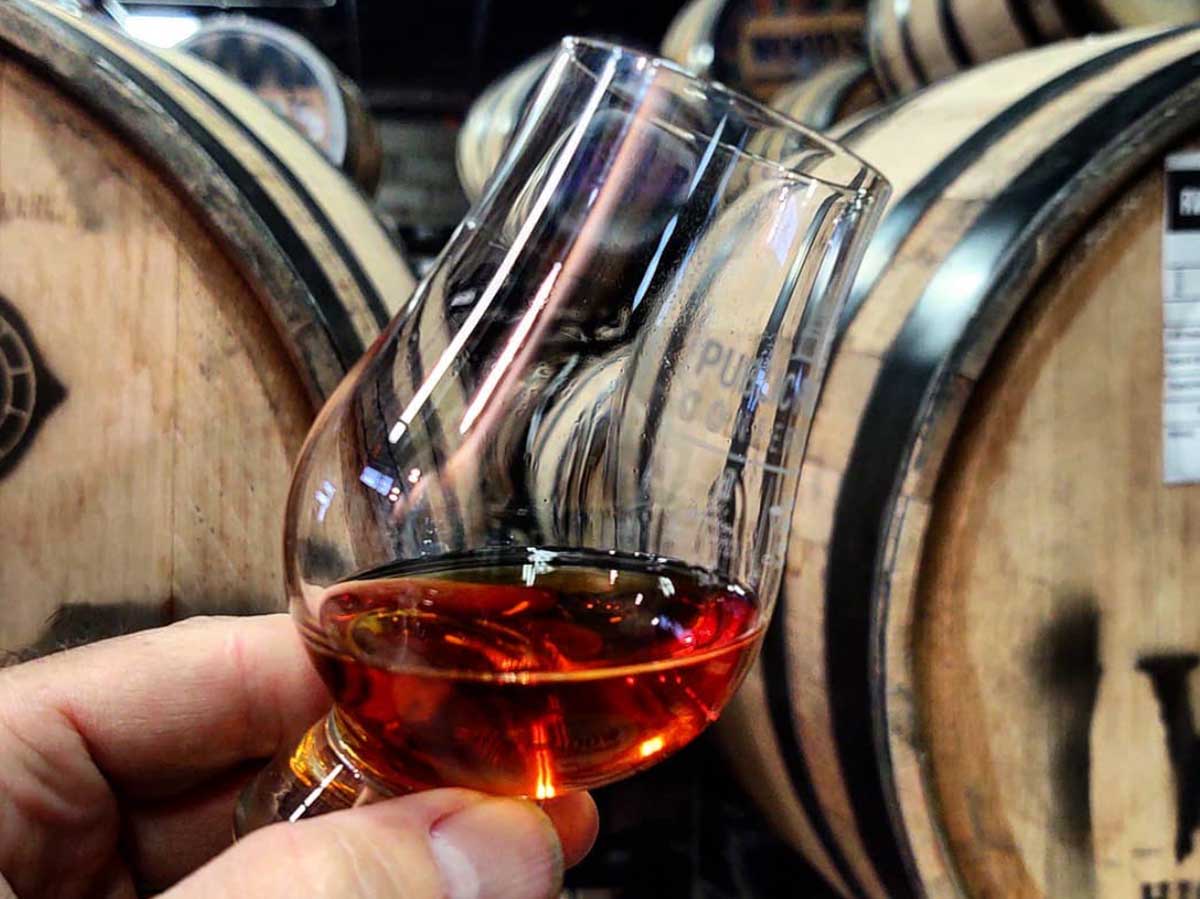 a glass of whiskey is held up next to a barrell at woods distillery