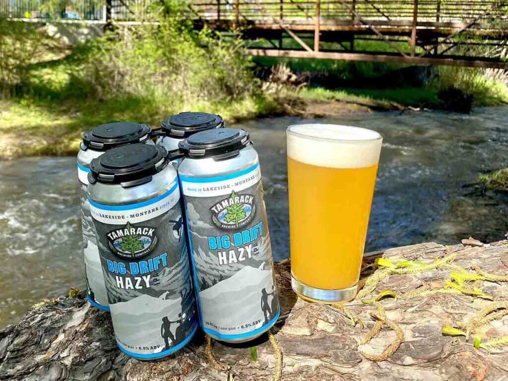 Tamarack Brewing Co. cans of beer, glass of beer set up on rock in front of a creek